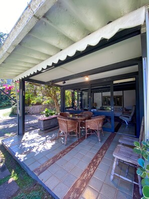 Una veranda esterna coperta con tavolo e sedie, un'area lounge con divano e poltrone, e vista sul giardino.