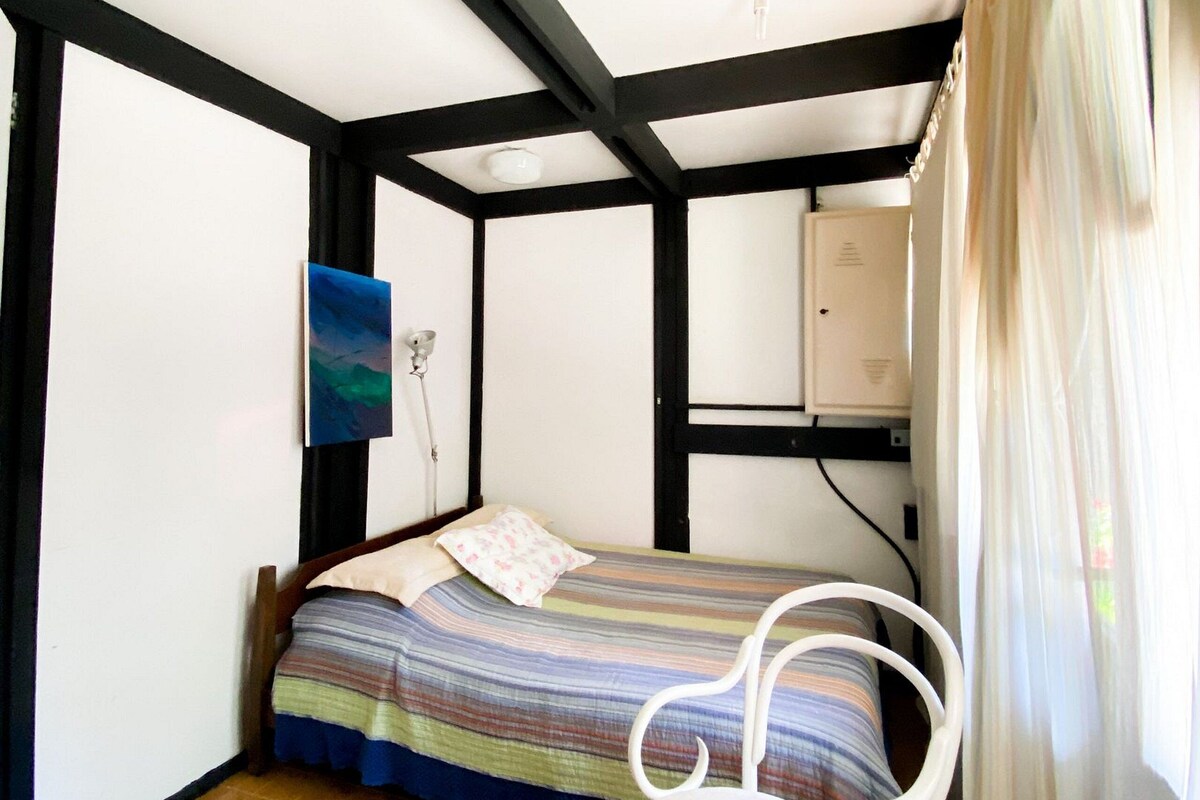 A small room with a bed, a chair, a painting on the wall, and a window with curtains.