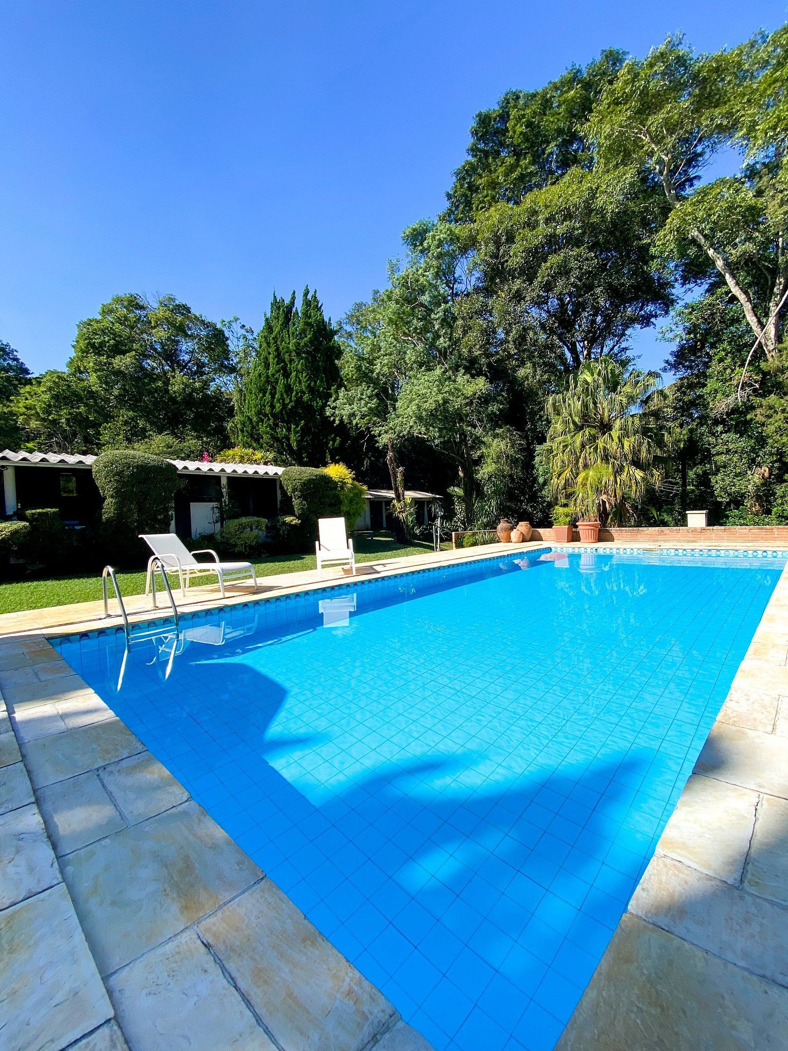 Piscina retangular com borda em azulejo, rodeada por um deck de pedra. Há espreguiçadeiras e um pequeno edifício com telhado em azulejo ao fundo.