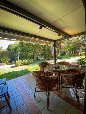 A covered patio with wicker furniture and a glass table, overlooking a well-maintained garden. - Chácara Espetacular em Ibiúna (Ibiuna)