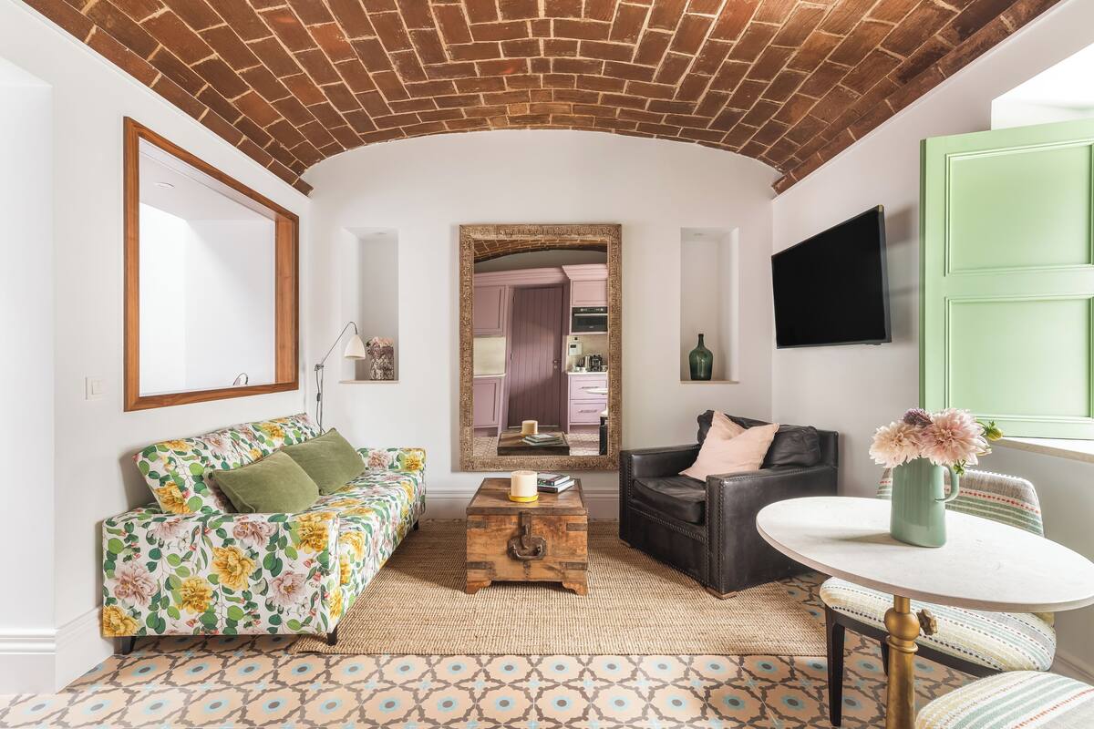 A living room with a brick ceiling, a sofa with floral bedding, a coffee table, a leather chair, a round table with a vase, and a mirror reflecting a kitchen.