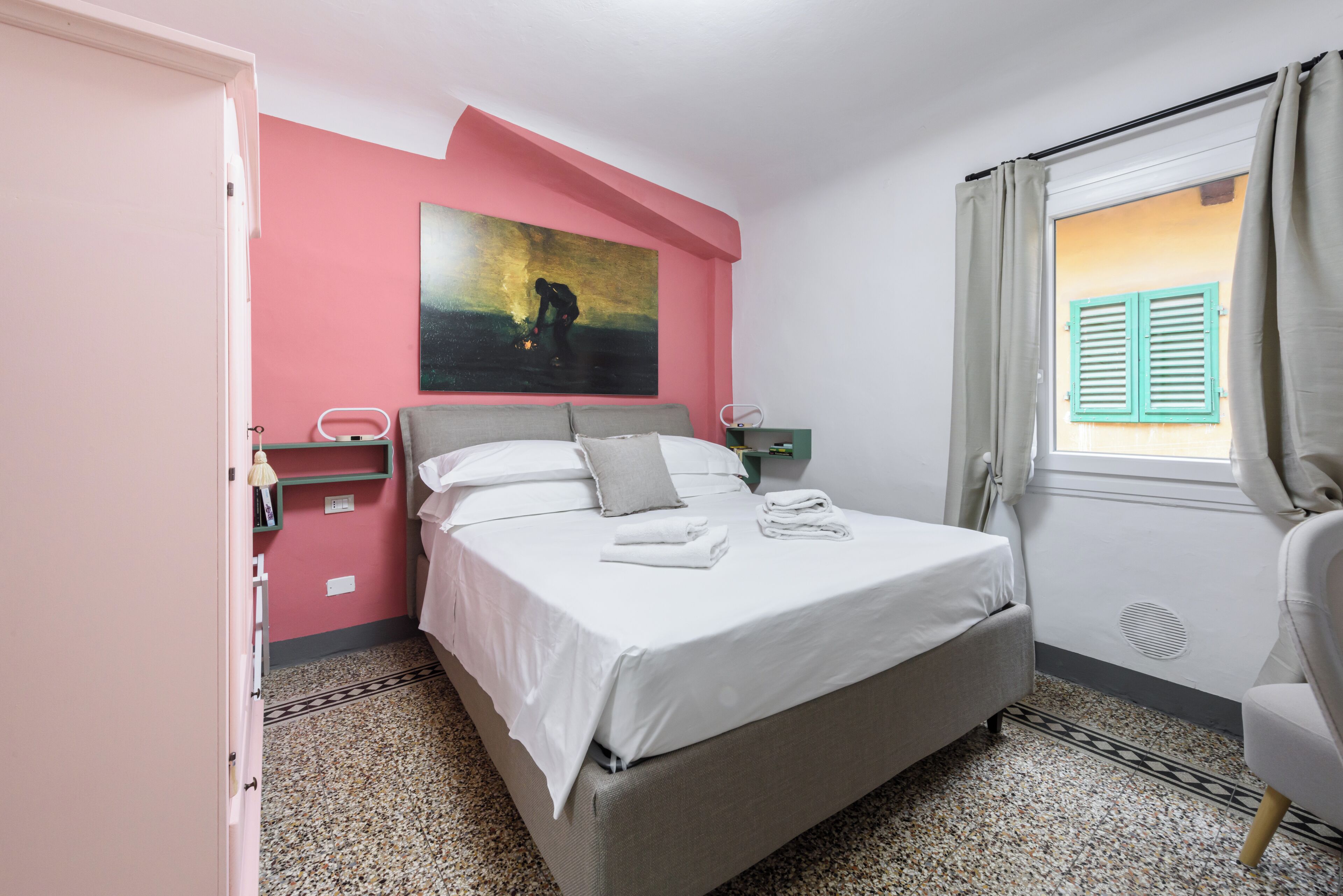 Ein Schlafzimmer mit Bett, einem Bild an der Wand, einem Fenster mit Vorhängen, einem Sessel und einem Nachttisch.