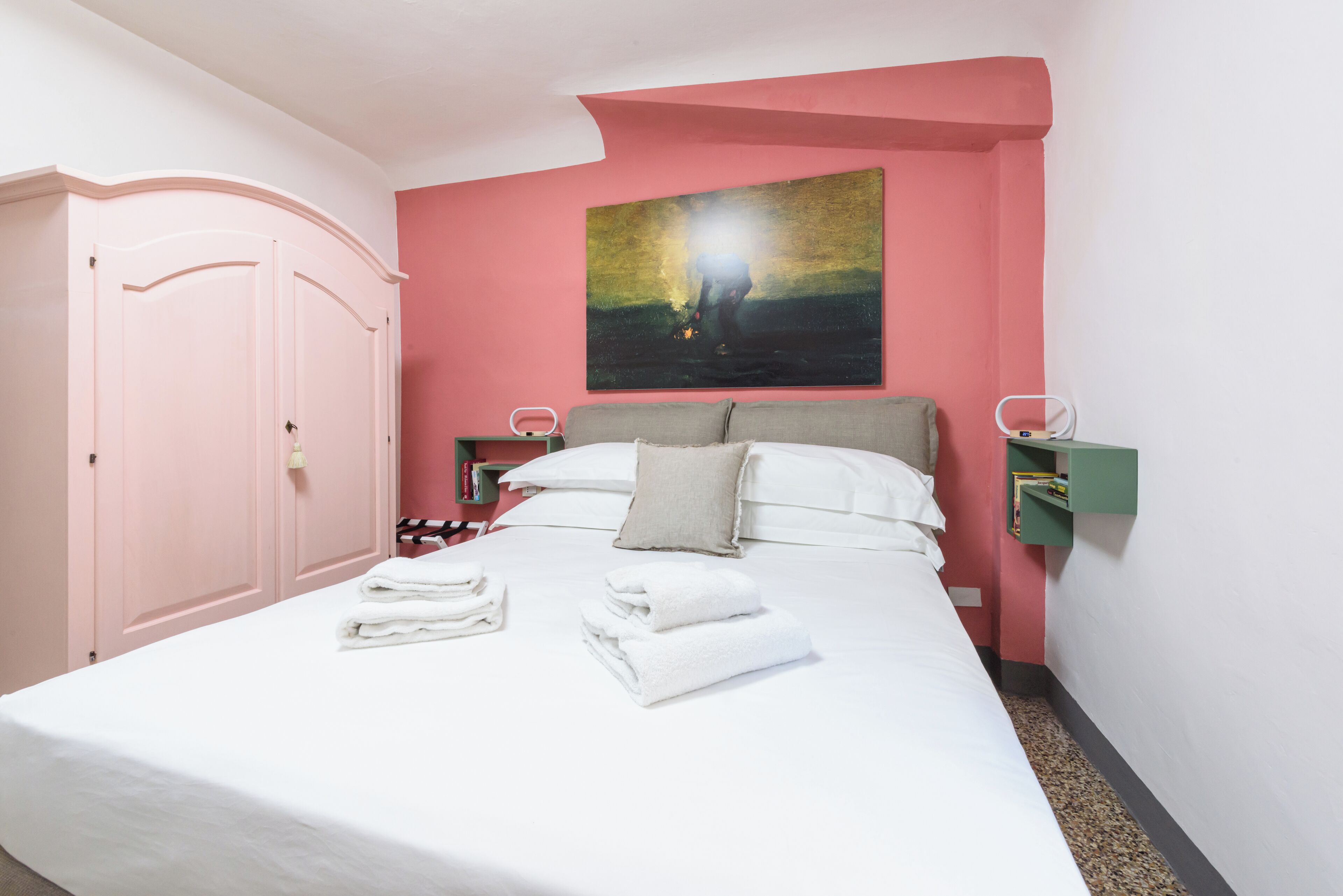 A neatly made bed with folded white towels, a pink wall, and a painting above it.