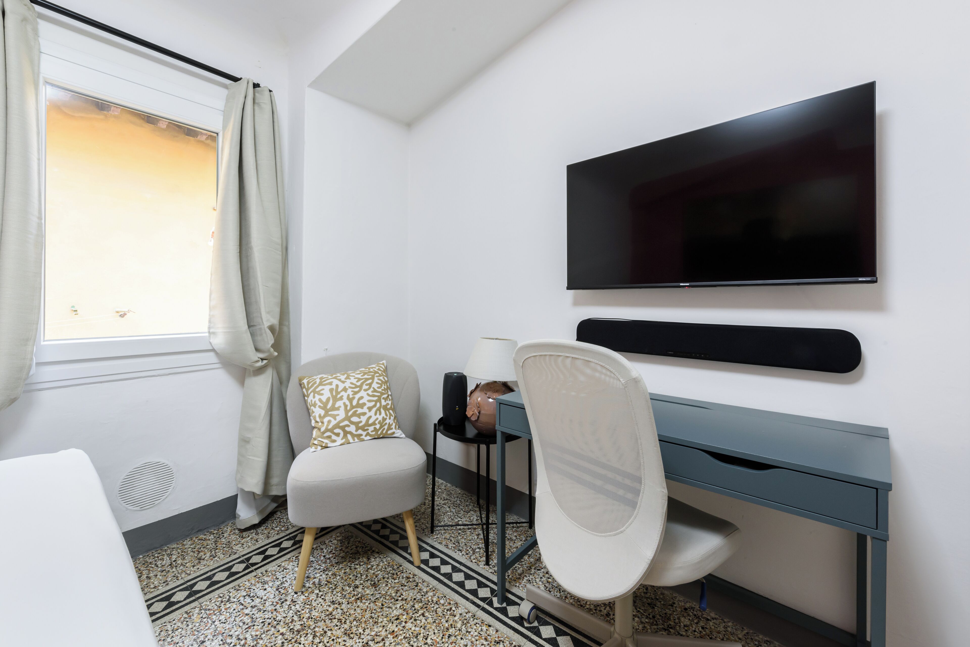A modern hotel room with a large flat-screen TV mounted on the wall, a desk with a lamp, a comfortable chair, and a patterned rug.