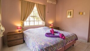 A bedroom with a bed, a nightstand, a window with curtains, and an air conditioning unit. - 03 Kas Koral by Bonaire Paradise (Kralendijk)