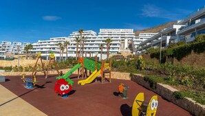 Un terrain de jeux doté de diverses installations, notamment un toboggan et des balançoires, situé devant un bâtiment moderne avec des balcons.