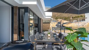 Un espace extérieur aménagé pour les repas, avec une table dressée pour quatre personnes, entourée de chaises, d’un barbecue et d’un grand parasol.
