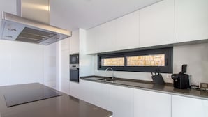Une cuisine moderne avec des armoires blanches, une hotte de cuisinière en acier inoxydable, une fenêtre intégrée avec un pare-feu en pierre, et une machine à café noire posée sur le plan de travail.