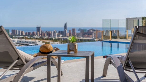 Un salon de bord de piscine avec vue sur la ville, une table agrémentée d’une plante et un chapeau de paille posé sur une chaise.