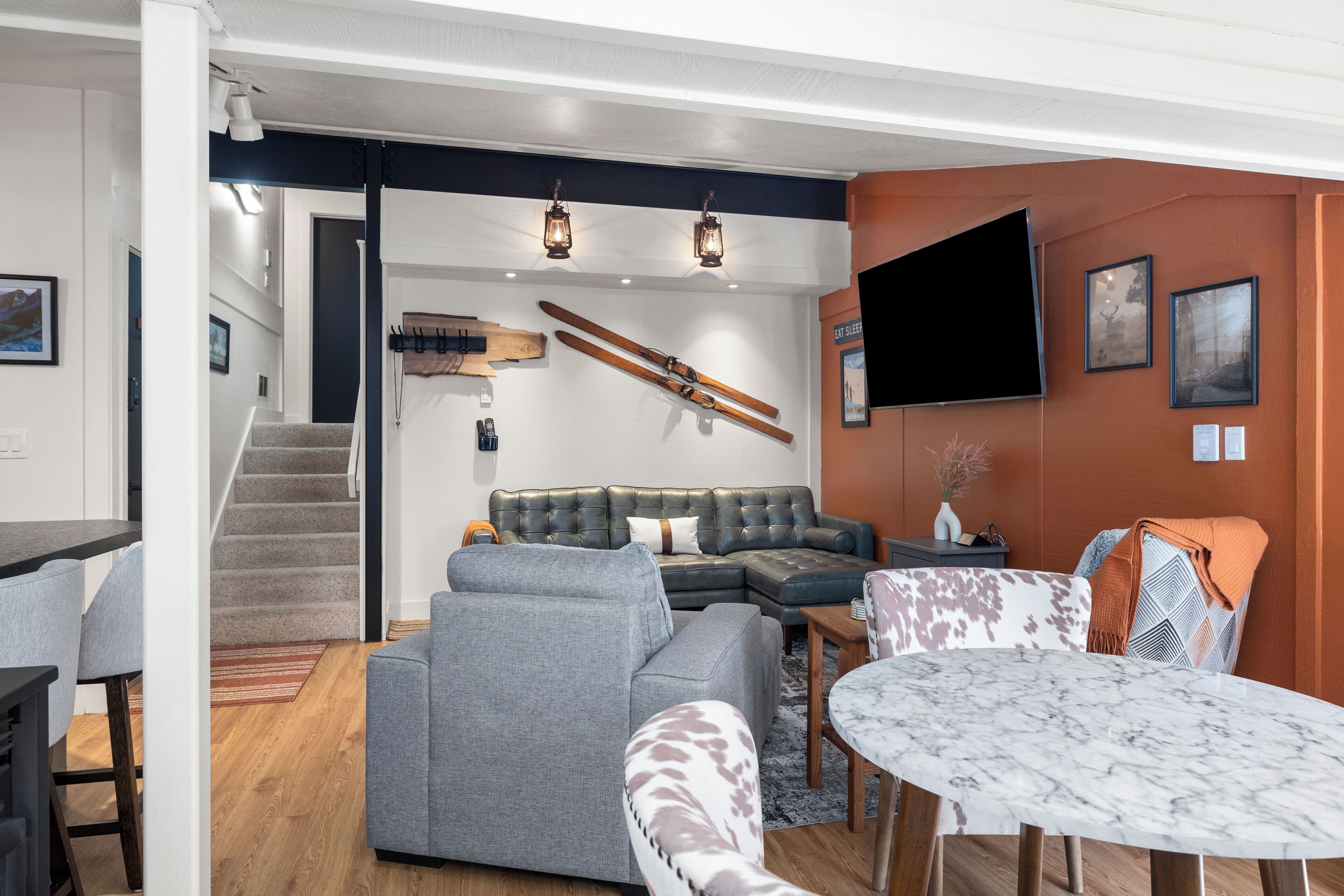 A modern living room with a grey sectional sofa, a coffee table, and a TV mounted on the wall. There is a staircase leading to another level, and a wooden bench with skis leaning against the wall.