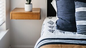Une chambre à coucher avec un lit, une table de chevet ornée d’une plante en pot, une lampe suspendue et une fenêtre avec des stores.