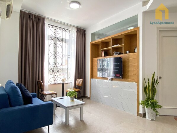 Un salon avec un canapé bleu, une petite table sur laquelle est posée une plante, un téléviseur fixé sur une étagère en bois et une plante en pot près de la porte.