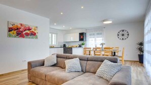 Un salon moderne avec un canapé d’angle, un coin repas avec une table et des chaises, une cuisine en arrière-plan et une horloge murale.