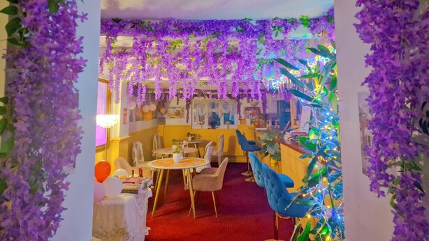 A restaurant with purple hanging flowers, round tables, and blue chairs. - The Evergreen hotel (Banbury)