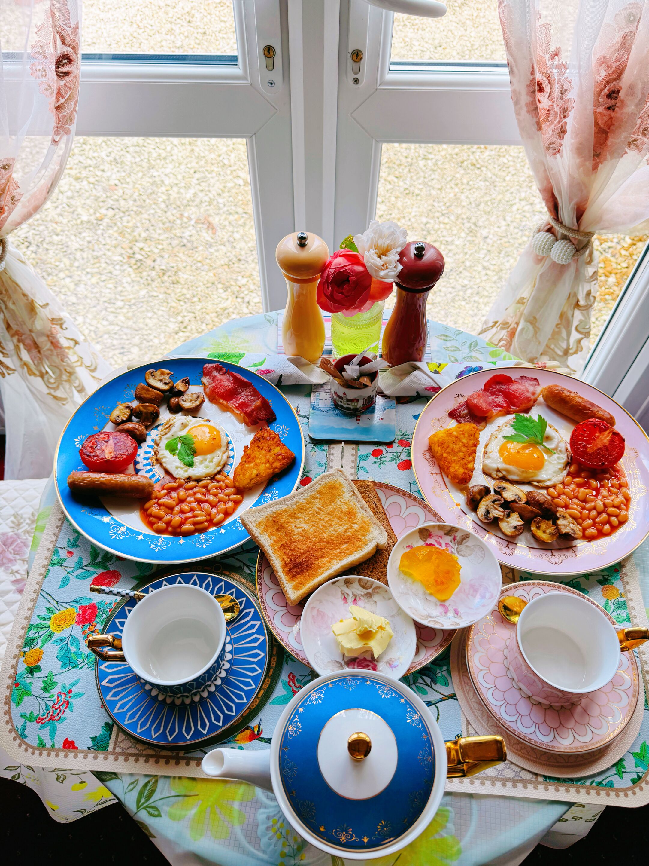Ein Frühstückstisch mit Tellern voller Eier, Würstchen, Bohnen, Toast und Marmelade. Salz- und Pfefferstreuer, ein Teekessel und Tassen sind ebenfalls vorhanden.