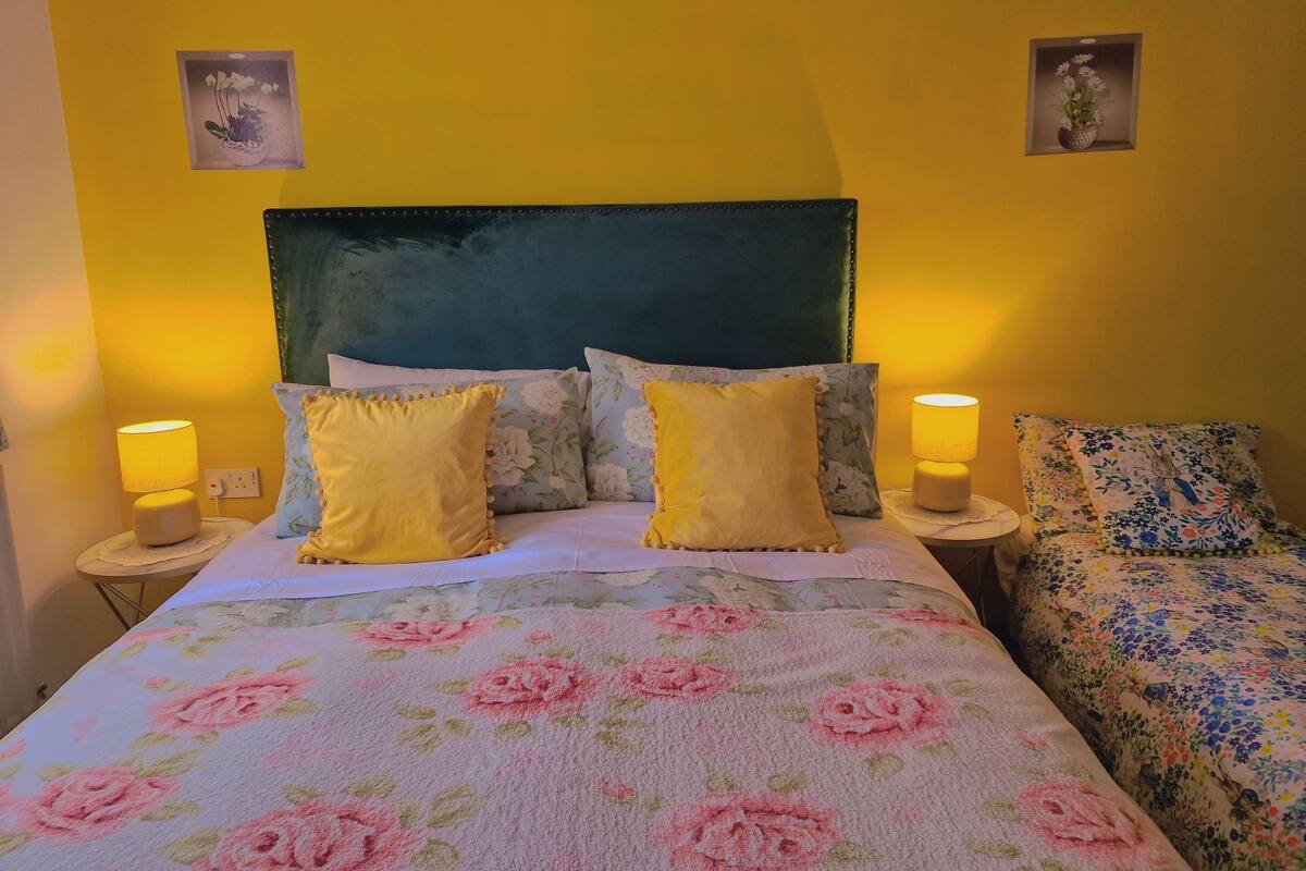 A bed with floral bedding, two bedside lamps, and a headboard against a yellow wall.
