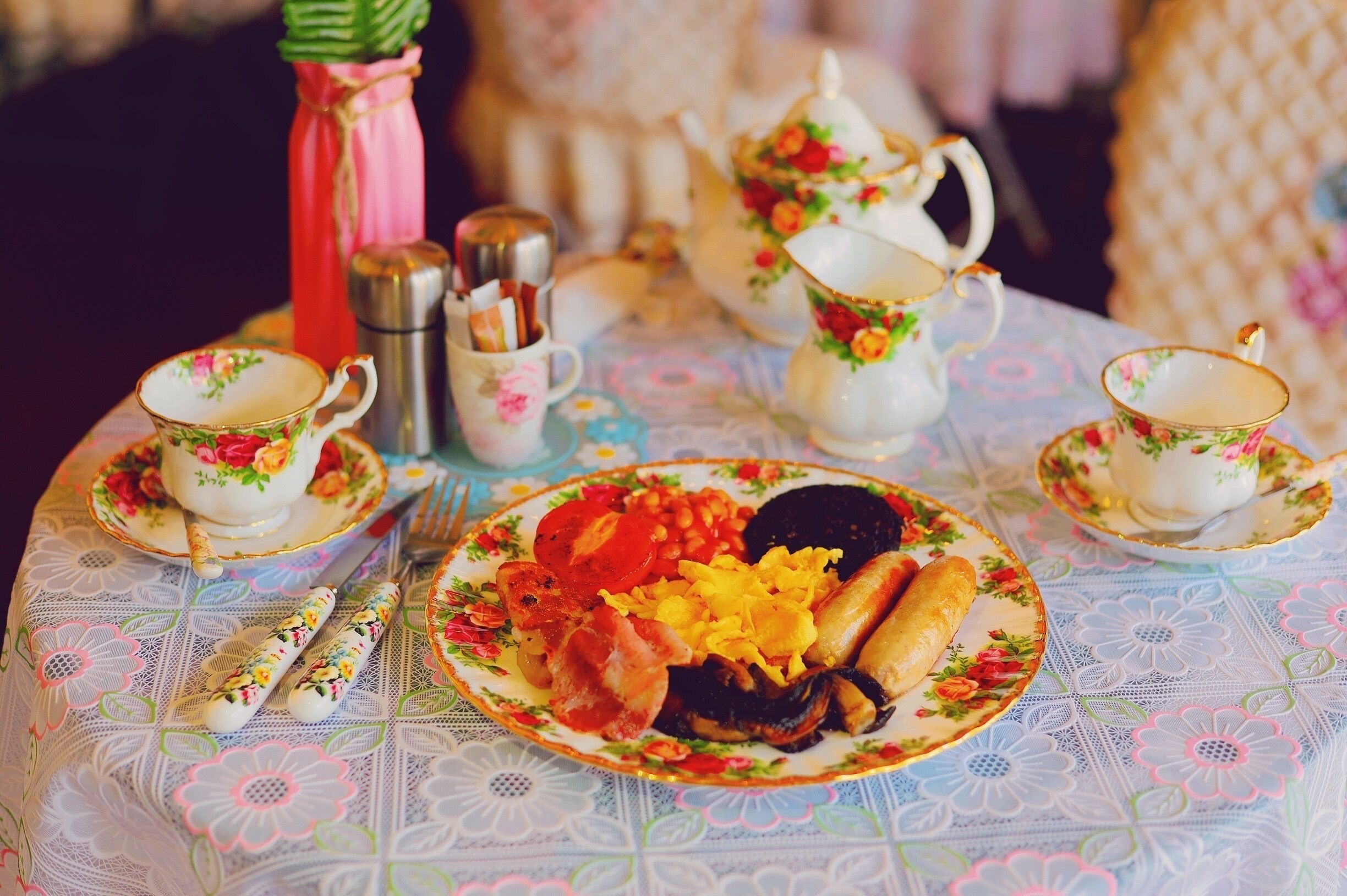 Una mesa dispuesta con vajilla de estampado floral, incluyendo tazas, platitos, una tetera y un azucarero, junto con un plato de comida.
