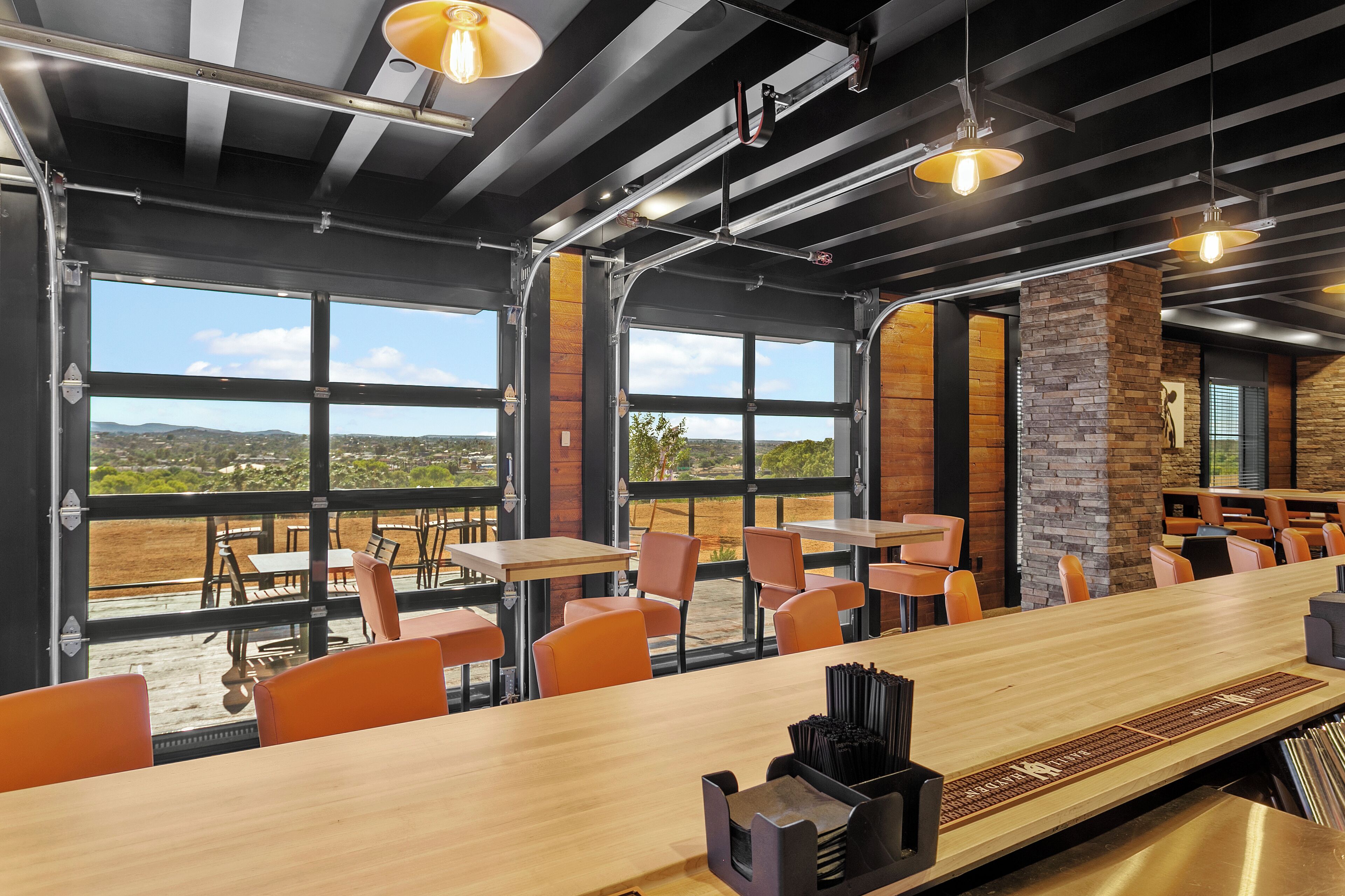 Un restaurante moderno con una barra de madera larga, sillas anaranjadas y amplias ventanas con vistas al exterior.