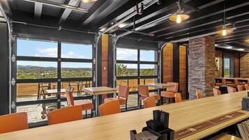 Un restaurante moderno con una barra de madera larga, sillas anaranjadas y amplias ventanas con vistas al exterior.