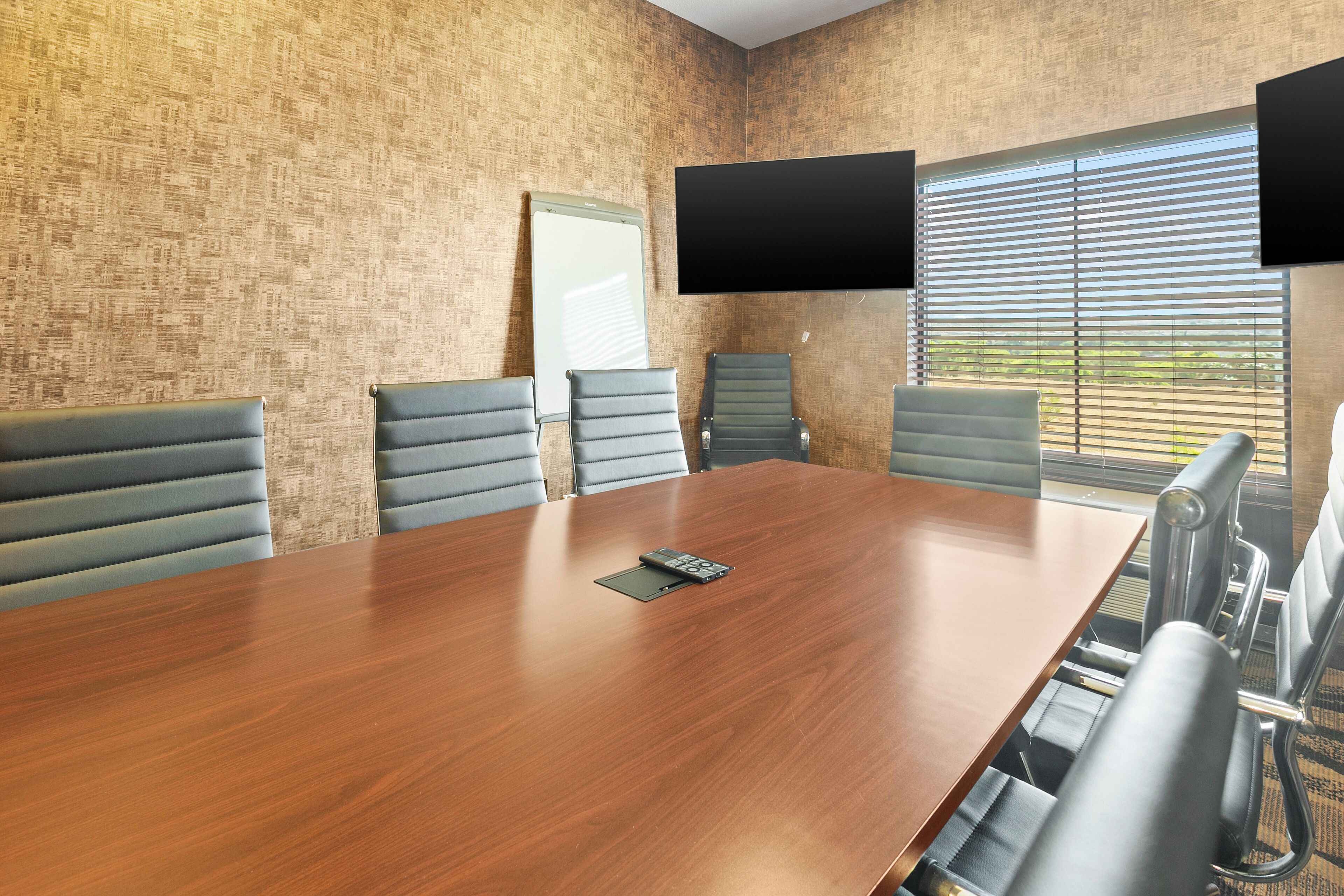 A conference room with a large wooden table, six grey upholstered chairs, a whiteboard, and a flat-screen TV mounted on the wall.