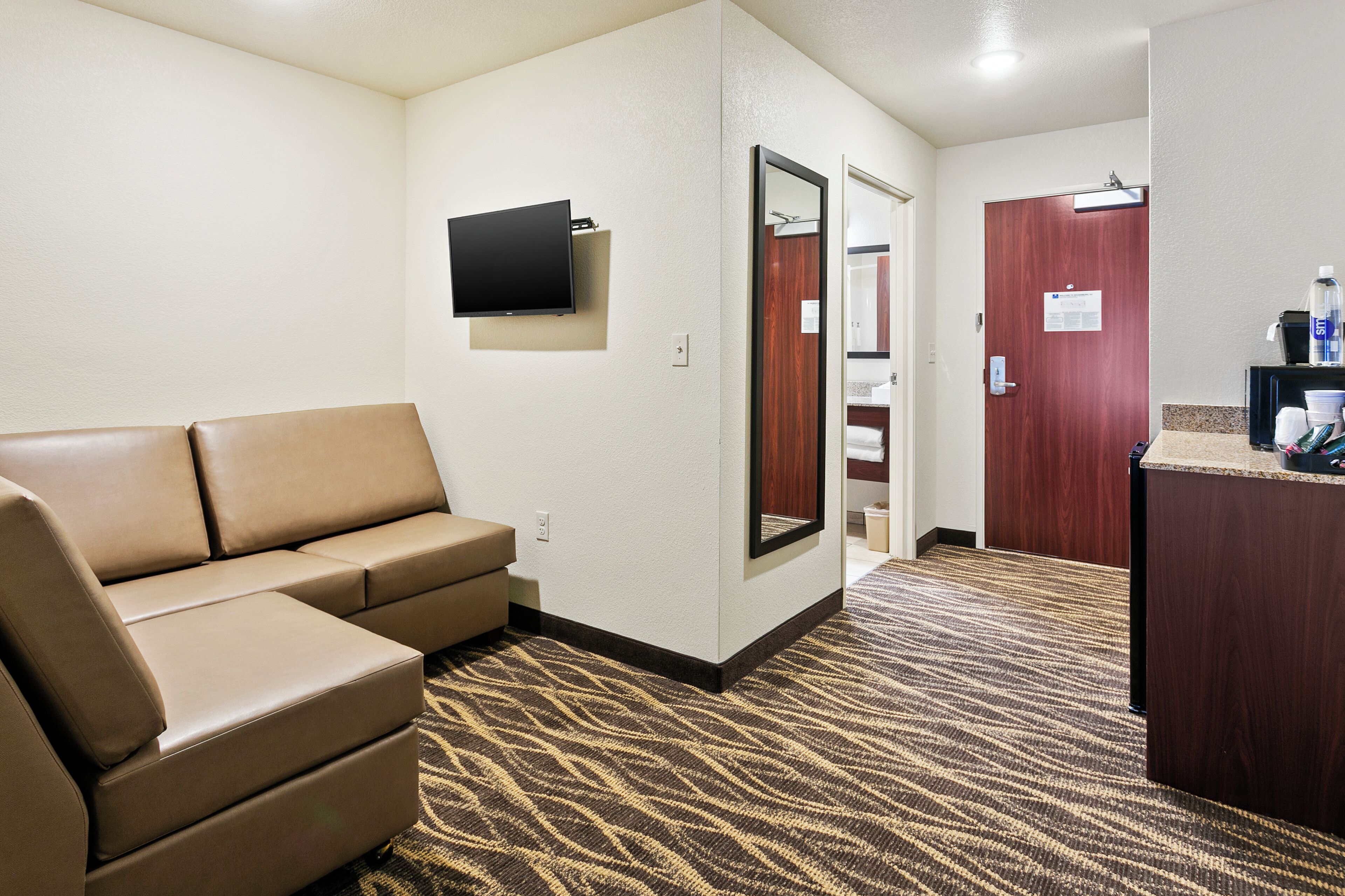 Habitación de hotel con sofá, televisor de pantalla plana, espejo, puerta de madera y un pequeño mostrador con artículos de aseo.