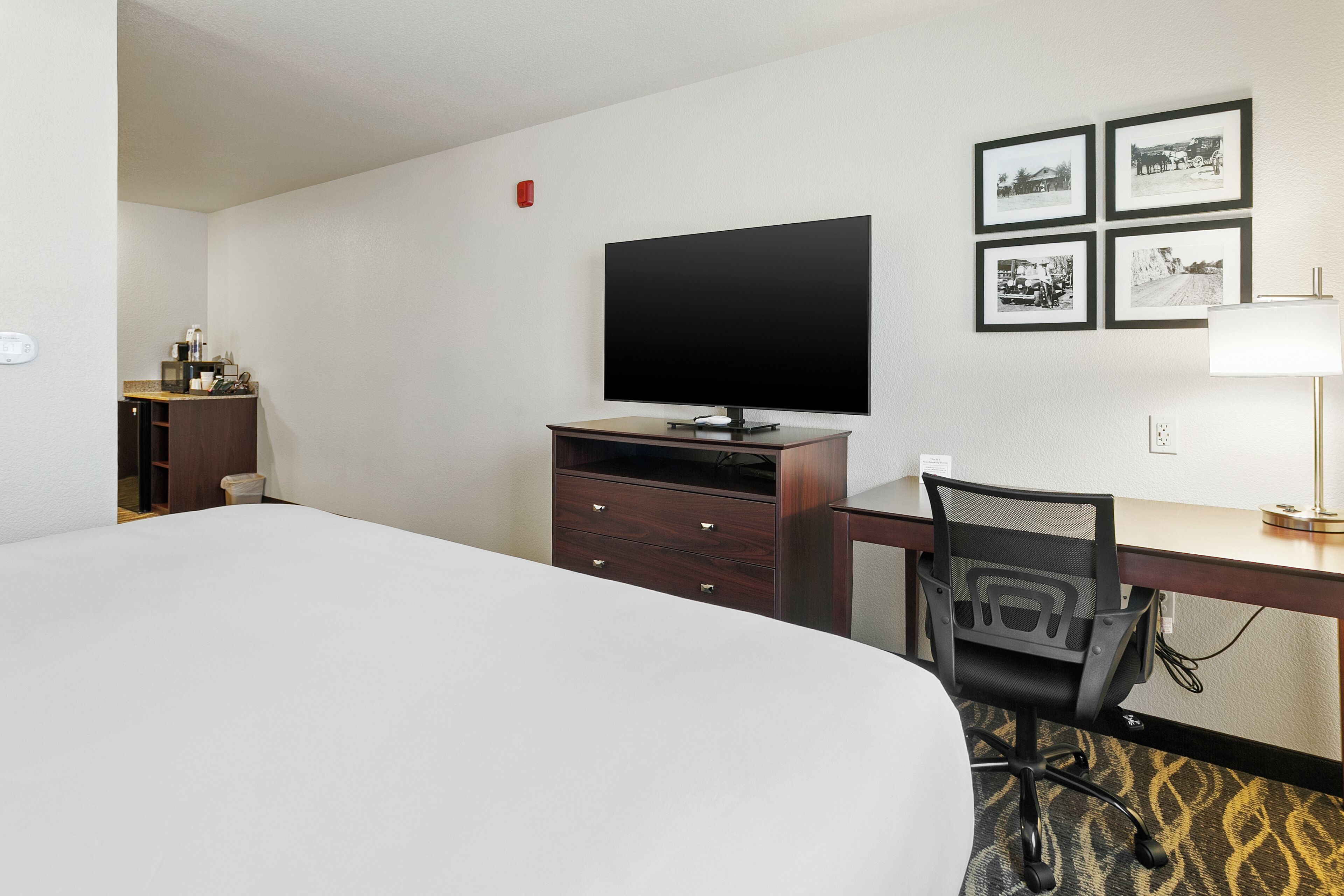 Habitación de hotel con una cama, un escritorio con silla, un televisor, un mueble pequeño y cuatro cuadros en la pared.