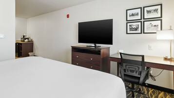 Habitación de hotel con una cama, un escritorio con silla, un televisor, un mueble pequeño y cuatro cuadros en la pared.