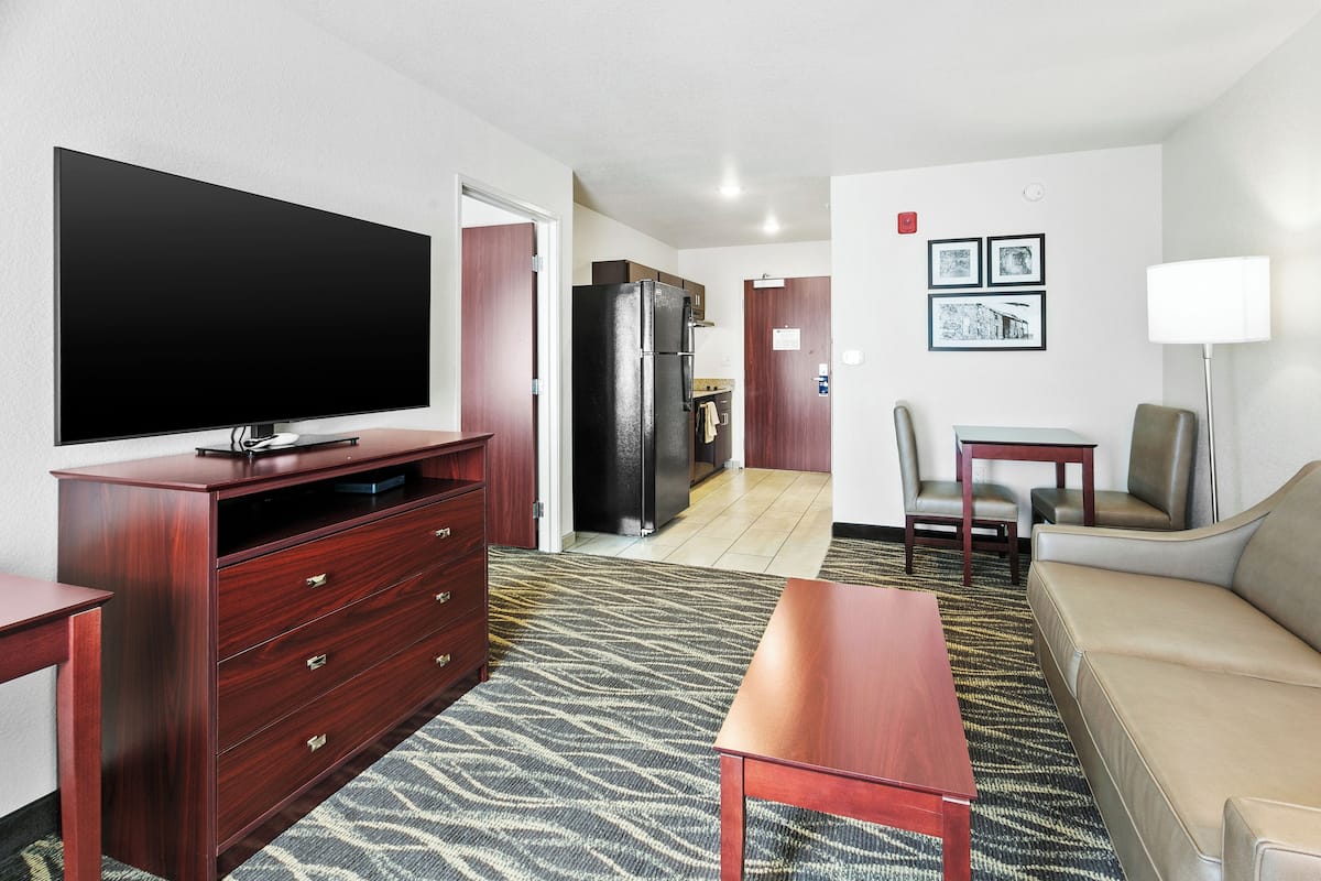 A hotel room with a flat-screen TV, wooden furniture, a sofa, a dining table, and a refrigerator.