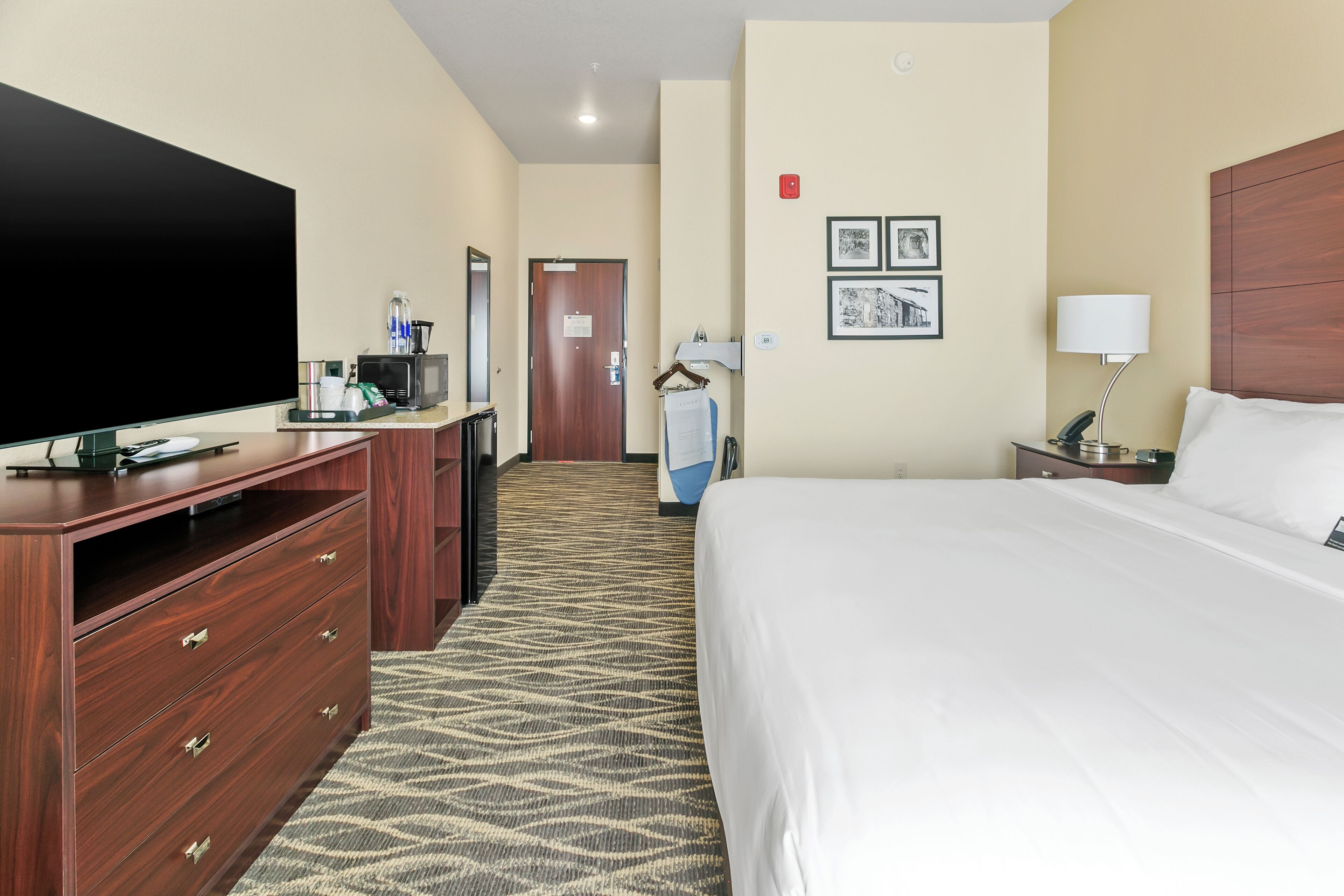 Hotelkamer met een groot bed, een televisie, een bureau met een computer, een koffiezetapparaat en een magnetron. Er hangen afbeeldingen in lijsten aan de muur en een deur is zichtbaar op de achtergrond.