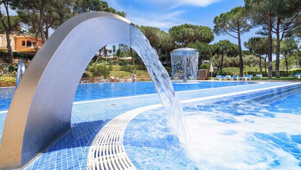 Una piscina con un moderno elemento acuático, rodeada de árboles y sillas de descanso.