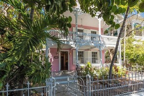 A two-story pink house with a balcony, surrounded by tropical plants and a white fence. - Historic Mansion in Old Town Key West with Private Pool - Sleeps 16! (Key West)