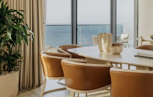A modern dining area with a large table and chairs, a potted plant, and a view of the sea through large windows.