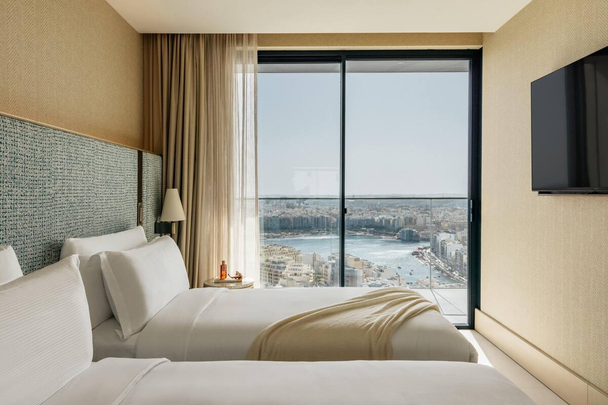 A hotel room with a large bed, a view of a cityscape through a floor-to-ceiling window, and a flat-screen TV mounted on the wall.