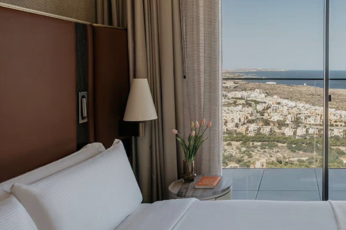 A hotel room with a bed, a lamp, a vase with flowers, and a view of a coastal cityscape.