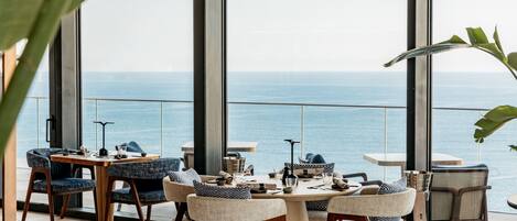 A modern dining area with large windows overlooking the sea, furnished with wooden chairs and tables.