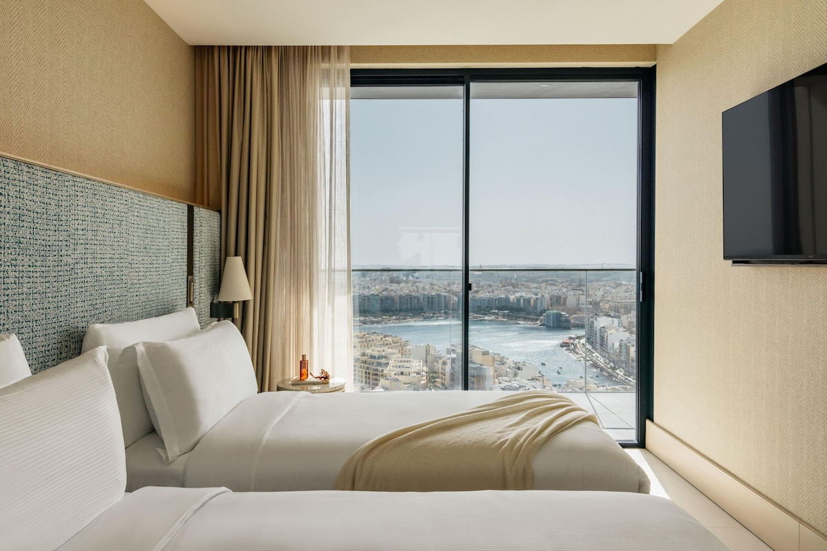 A hotel room with a large bed, a view of a cityscape through a floor-to-ceiling window, and a flat-screen TV mounted on the wall.