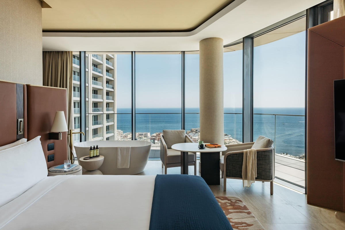 A hotel room with a large bed, a sitting area with a view of the sea, a bathtub, and a television.