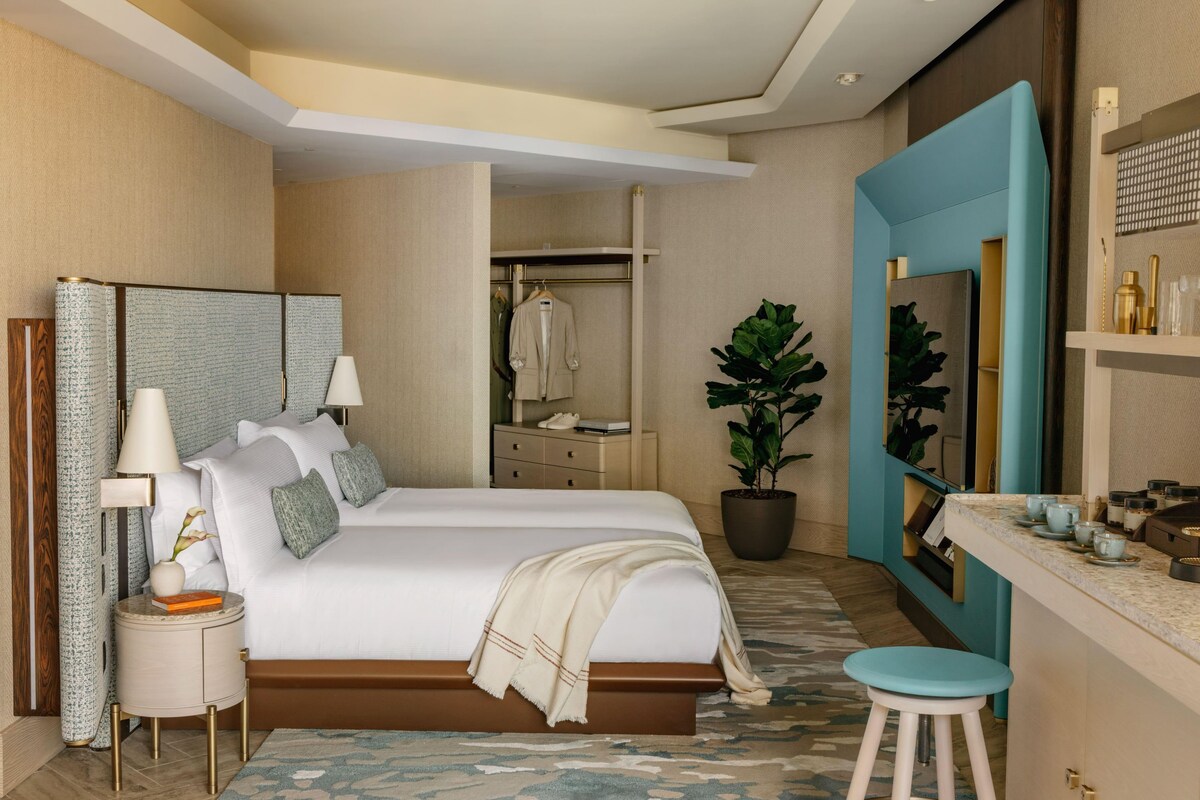 Una habitación de hotel moderna con cama, mesita de noche, lámpara, planta en maceta y baño con espejo y estante.