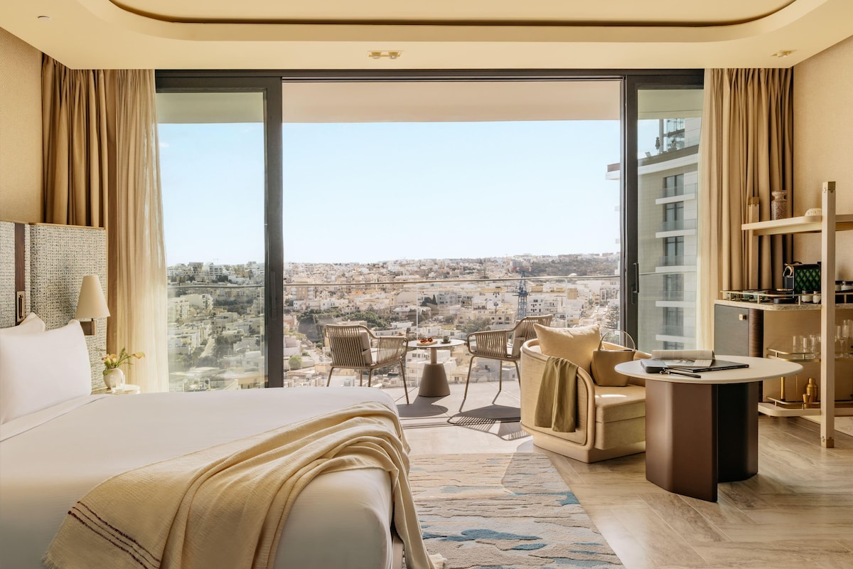 A modern hotel room with a large bed, a seating area with a table, and a view of the city.