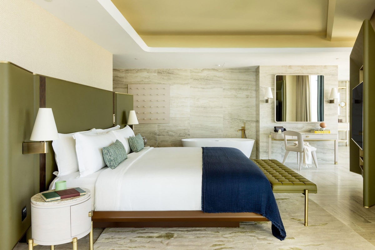 A modern hotel room with a large bed, bedside tables, a vanity with a mirror, and a seating area.