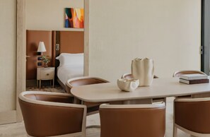 Una habitación de hotel con cama, una mesita de noche con lámpara, una planta pequeña, un cuadro en la pared y una zona de comedor con sillas y mesa.
