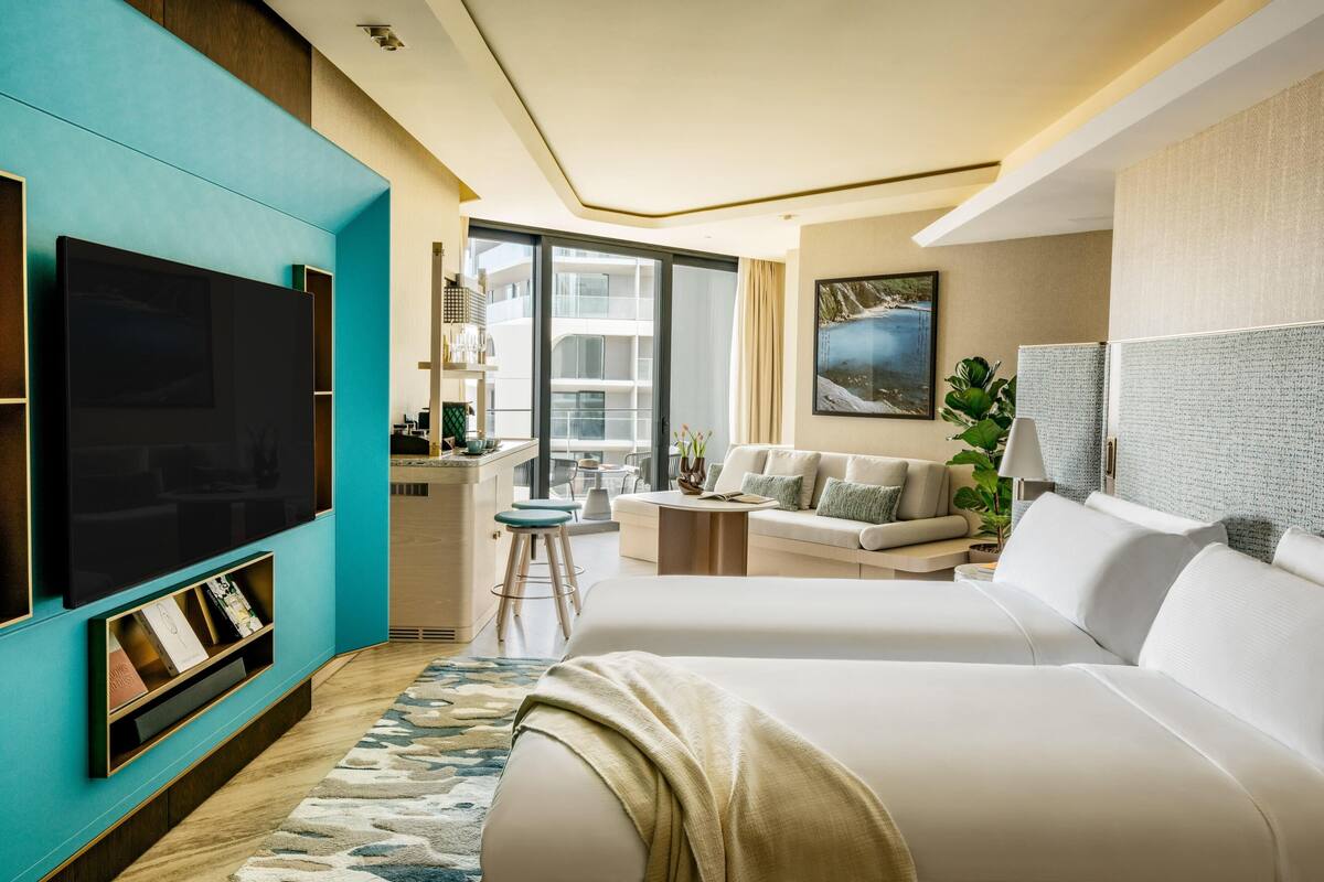 A modern hotel room with a large flat-screen TV, a sofa, a bed, a small kitchen area, and a balcony with a view.