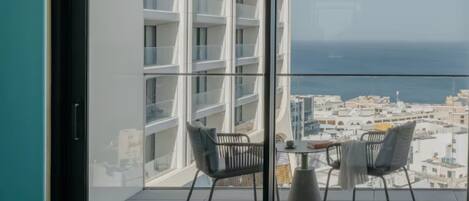 A balcony with a table and chairs, a bed, and a view of a cityscape and sea.