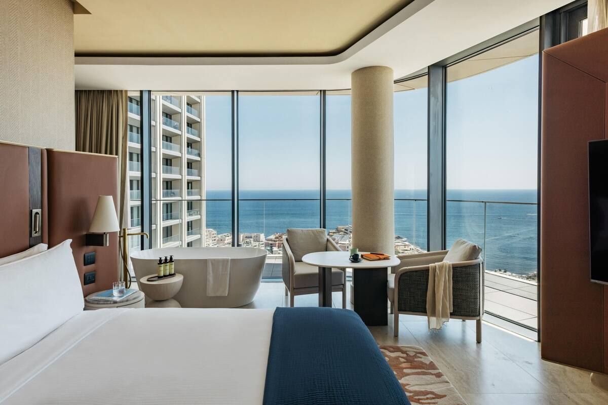 A hotel room with a large bed, a sitting area with a view of the sea, a bathtub, and a television.