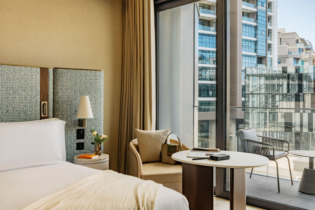 A hotel room with a large bed, a round table, a chair, a sofa, a lamp, a nightstand, and a view of the city through large windows.