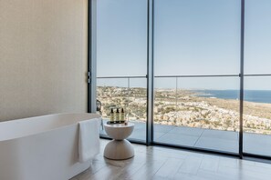 Un baño moderno con bañera, una mesa redonda de madera con artículos de aseo, y un ventanal con vistas a la ciudad.