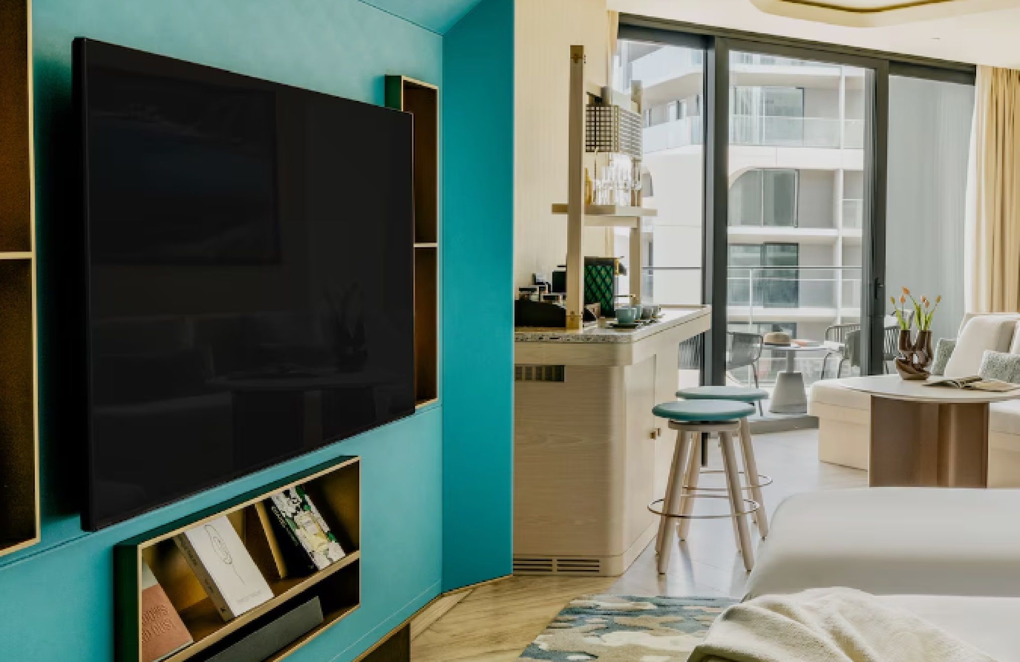 Une chambre d’hôtel moderne dotée d’une grande télévision à écran plat, d’un balcon avec un coin repas et offrant une vue sur la façade de l’immeuble.