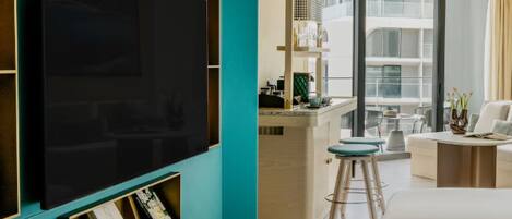 Una habitación de hotel moderna con un televisor de pantalla plana grande, un balcón con zona de comedor y vistas del exterior del edificio.