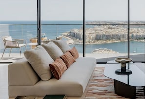 A modern living room with a large window, a sofa, a coffee table, and a view of a coastal cityscape.