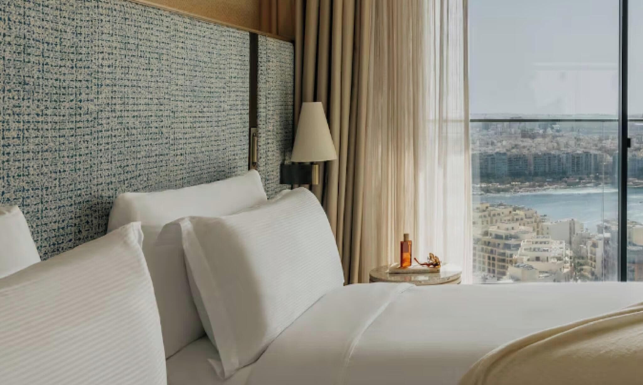 Quarto de hotel com uma cama grande, uma mesa de cabeceira, uma luminária e vista para a cidade através de uma janela ampla.